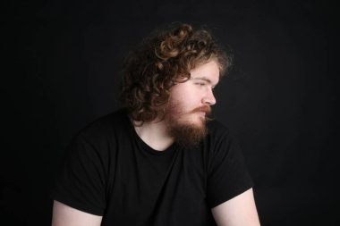 Plus size male with long hair and a beard doing a sultry pose in front of a studio black backdrop  