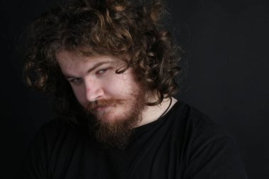 Plus size male with long hair and a beard doing a sultry pose in front of a studio black backdrop  