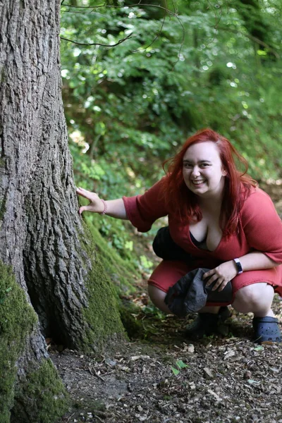 Plus size female crouched down by a tree in the forest looking for a fairy house in a hole