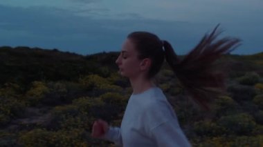 Güzel bir kız akşamleyin yolda koşar. Yüksek kalite 4K görüntü. Büyük çekim, tepenin arkasına, çimenlere ve akşam gökyüzüne karşı. Kız beyaz bir kazak giyiyor..