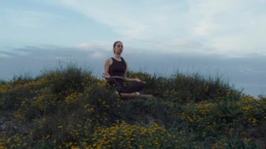Genç bir atletik kız deniz kenarındaki bir tepede lotus pozisyonunda meditasyon yapıyor. Yüksek kalite 4K görüntü. Kız sarı çiçeklerin arasında oturuyor. Arkaplanda bulutlu bir günbatımı gökyüzü var..