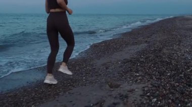 Akşamüstü gökyüzünün arka planında deniz kenarında kumlu bir sahil boyunca koşan sportif takım elbiseli genç ve atletik bir kızın orta boy çekimi.. 