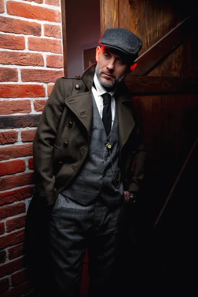 A man in a classic suit and coat stands on the stairs near the house, looking forward with wariness. There is danger ahead.