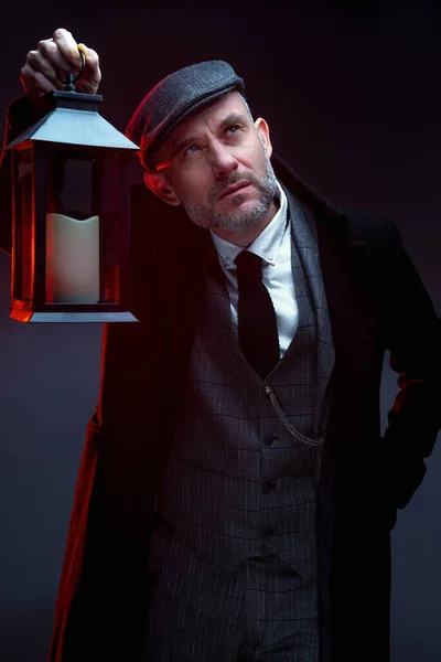 A man in a classic suit, coat and cap. Stands. Holds a vintage lantern in his hand. Upward gaze. Studio photography.