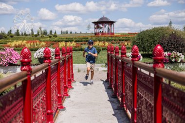 Şapkalı çocuk parktaki köprü boyunca koşuyor.