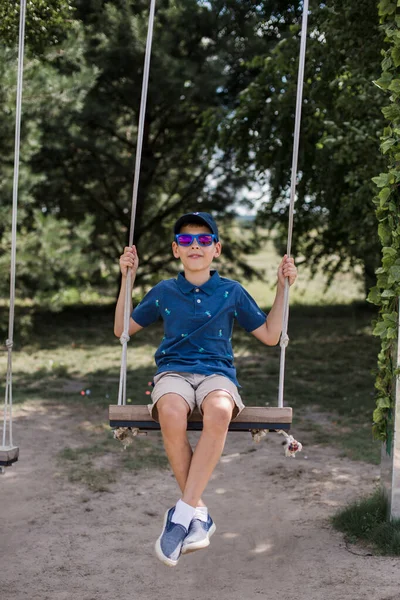 Güneş gözlüklü çocuk yazın parkta salıncakta sallanıyor.