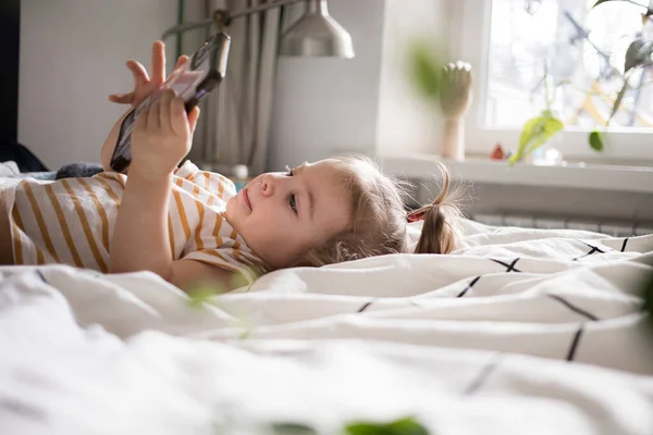 Klein meisje dat thuis met een smartphone op het bed speelt. blank peuter meisje. — Stockfoto