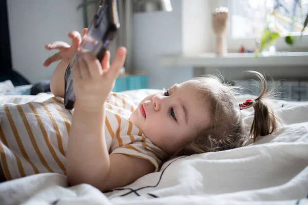 Klein meisje dat thuis met een smartphone op het bed speelt. blank peuter meisje. — Stockfoto