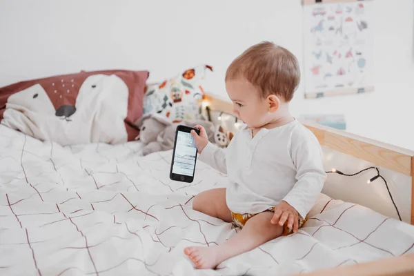 Baby meisje zit op bed en het gebruik van de smartphone in neutrale tinten kinderkamer. Geslacht neutrale babykamer en kleding — Stockfoto