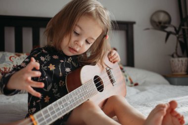 Ukelele gitar çalan küçük bir kız evdeki yatakta oturuyor.