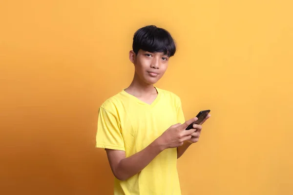 Portrait of Asian young man wearing yellow t-shirt looking happy and seeing news from smart phone, looking at the camera. Half body portrait on yellow background.