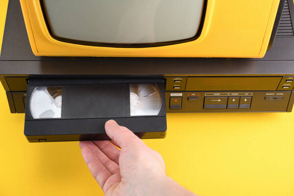Видеокассету VHS помещают в видеомагнитофон для просмотра видео. Old yellow vintage TV with VCR and videotape on black background from 1980, 1990, 2000. 