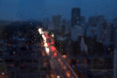 Bulanık pencere. Bardaktaki yağmur damlaları. Şehir yukarıdan görünüyor. Renkli bokeh, trafik ışıkları, binalar ve mavi gökyüzü. Paulo, Brezilya