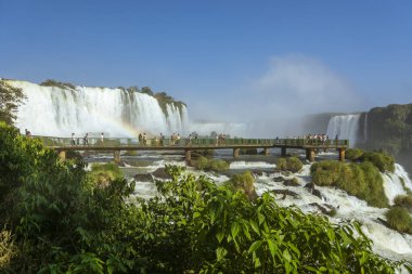 Foz do Iguacu, Parana, Brezilya. Güneşli bir günde Iguazu Şelalesi. Park yollarında ziyaretçiler var. Gökkuşağıyla güçlü bir su akıntısı.