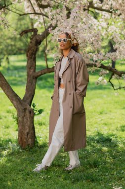 young african american woman in stylish sunglasses and trench coat walking with hands in pockets in park