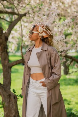young african american woman in stylish sunglasses and trench coat standing with hand in pocket in park