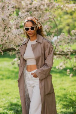 young african american woman in stylish sunglasses and trench coat standing with hand in pocket near blooming tree