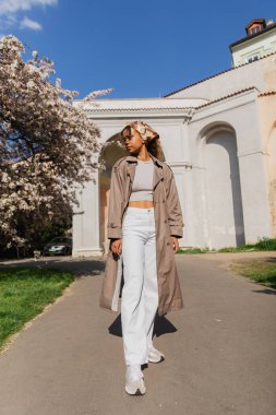 full length of stylish african american woman in trench coat standing in park near blooming tree 