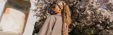 young african american woman in headscarf and stylish trench coat standing near blooming tree in prague, banner