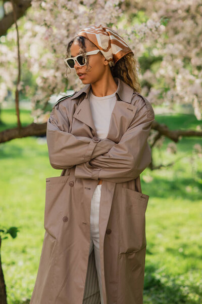 young african american woman in stylish sunglasses and trench coat standing with crossed arms near blooming tree