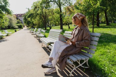 african american woman in headscarf and stylish trench coat sitting on bench and reading magazine in park