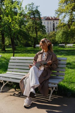 young african american woman in headscarf and trendy outfit sitting on bench and reading magazine
