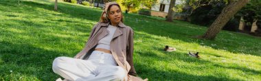 young african american woman in headscarf and stylish trench coat resting on green grass in park, banner clipart
