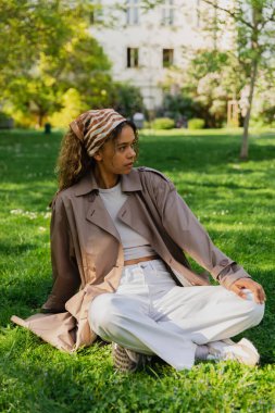 stylish african american woman in headscarf and trench coat sitting on green lawn in park