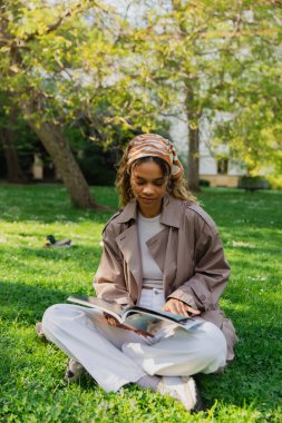 african american woman in headscarf and trench coat sitting on green lawn and reading magazine in park