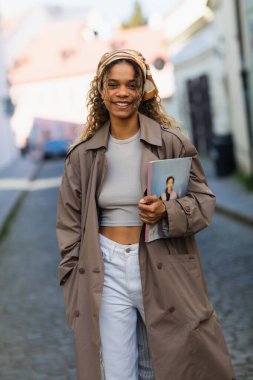 happy african american woman in headscarf and trench coat walking with magazine on street in prague 