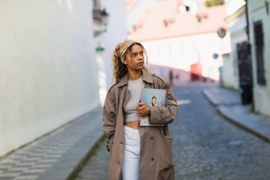 stylish african american woman in headscarf and trench coat walking with magazine on street in prague 