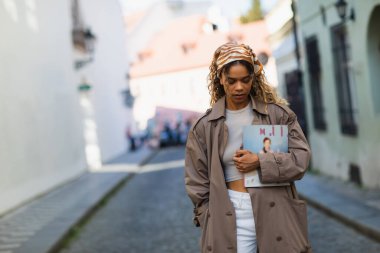 curly african american woman in headscarf and trench coat walking with magazine on street in prague 
