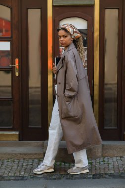 full length of stylish african american woman in headscarf and trench coat walking with hand in pocket in prague