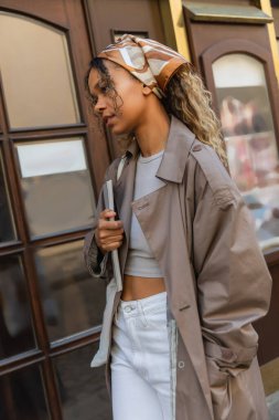 stylish african american woman in headscarf and trench coat walking with hand in pocket in prague