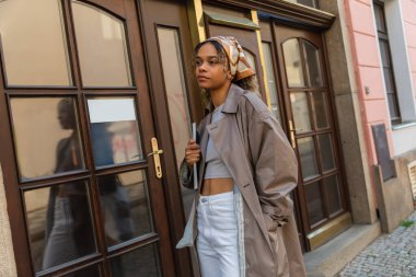 stylish african american woman in headscarf and trench coat standing with hand in pocket near entrance door in prague