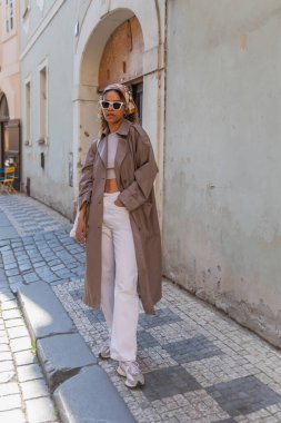 full length of stylish african american woman in headscarf and sunglasses posing in trench coat on street in prague 