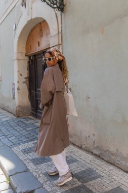 full length of stylish african american woman in trendy accessories posing with reusable bag on street in prague