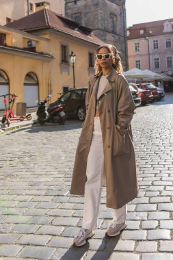 full length of curly african american woman in stylish accessories posing with hand in pocket on street in prague