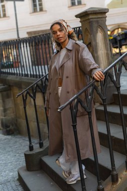 full length of trendy african american woman in trench coat standing on stairs 
