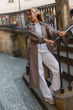 full length of amazed african american woman in trench coat standing on stairs 