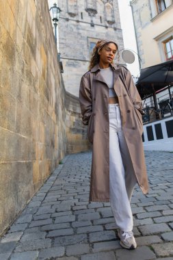 full length of stylish african american woman in trendy trench coat walking with hands in pockets near ancient wall in prague