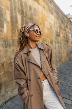 stylish african american woman in trendy accessories and trench coat listening music in wired earphones 