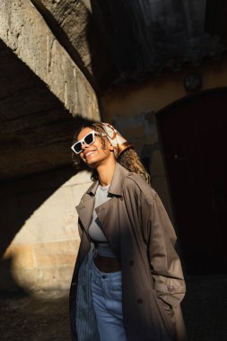 happy african american woman in trendy accessories and trench coat standing on spotlight from sunlight 