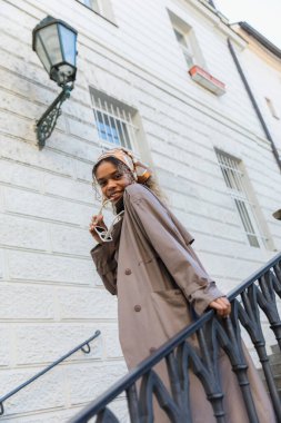 low angle view of happy african american woman in trendy headscarf holding sunglasses and standing on stairs in prague 