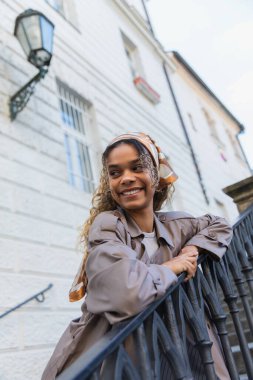cheerful african american woman in headscarf and trendy trench coat touching handrail while standing on stairs in prague 