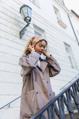 african american woman in trench coat adjusting headscarf on street in prague