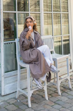 african american woman in stylish trench coat eating ice cream and sitting on chair in prague