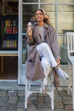 cheerful african american woman in headscarf and stylish trench coat holding ice cream cone and sitting on chair in prague
