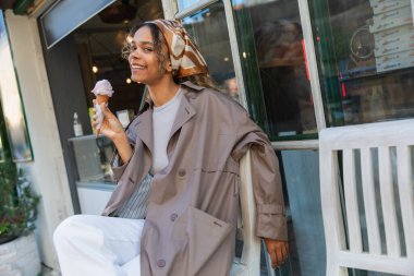 pleased african american woman in headscarf and stylish trench coat holding ice cream cone and sitting on chair in prague