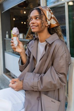 cheerful african american woman in headscarf holding ice cream cone and sitting on chair in prague